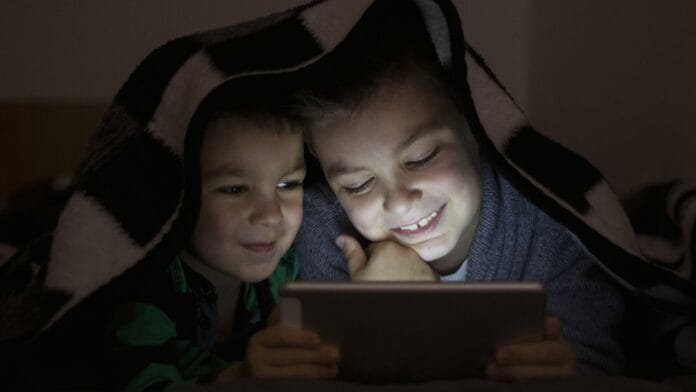 ninos-usando-tablet-noche-tendencias-digitales-2025 Dos niños usando tablet bajo una manta en la oscuridad, representando el tiempo de pantalla nocturno y las tendencias digitales infantiles en 2025